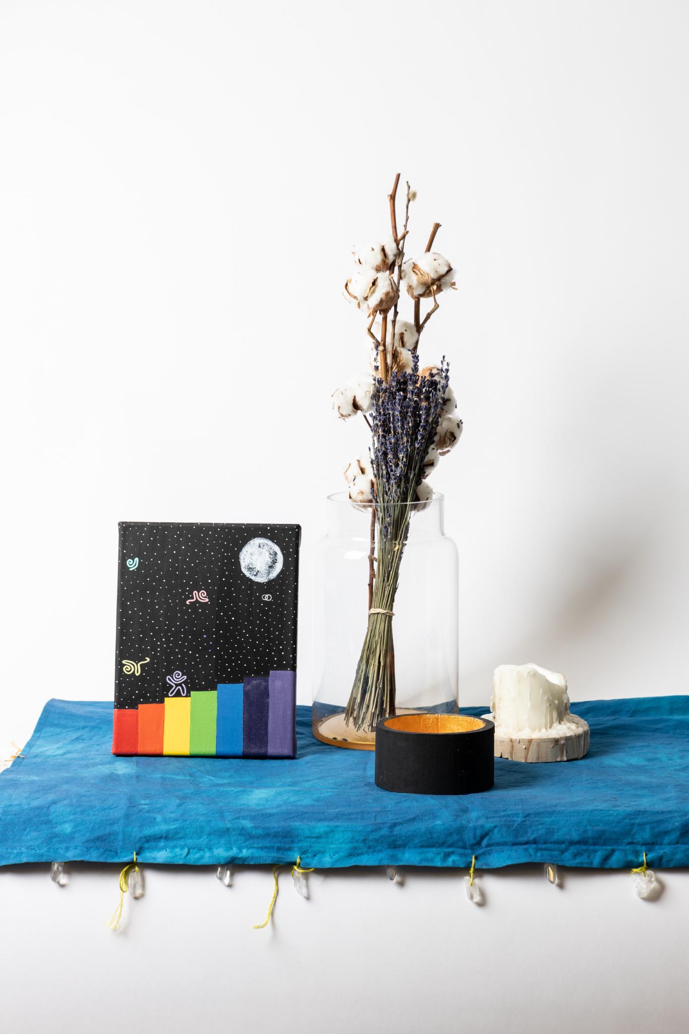 A painting, dried flowers, black bowl, and candle clustered on a blue fabric square.