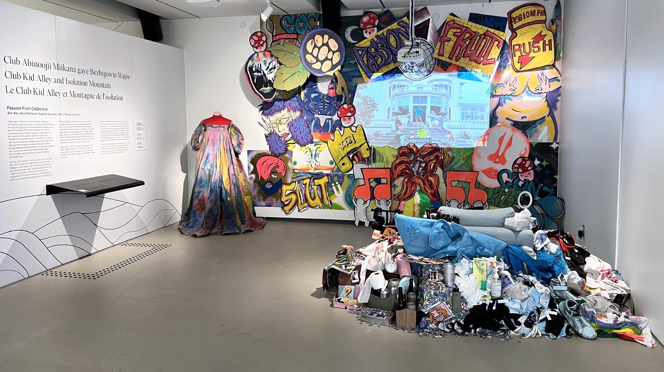 A bright installation made out of objects mounded around a small chair facing a video projection framed by neon graffiti cardboard cut outs and a colorful gown in the left corner.