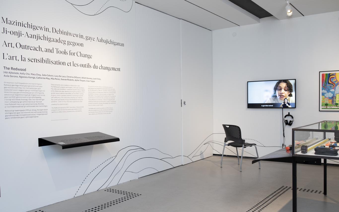 A corner of a white gallery with a TV, framed painting, and large glass case set away from the wall.