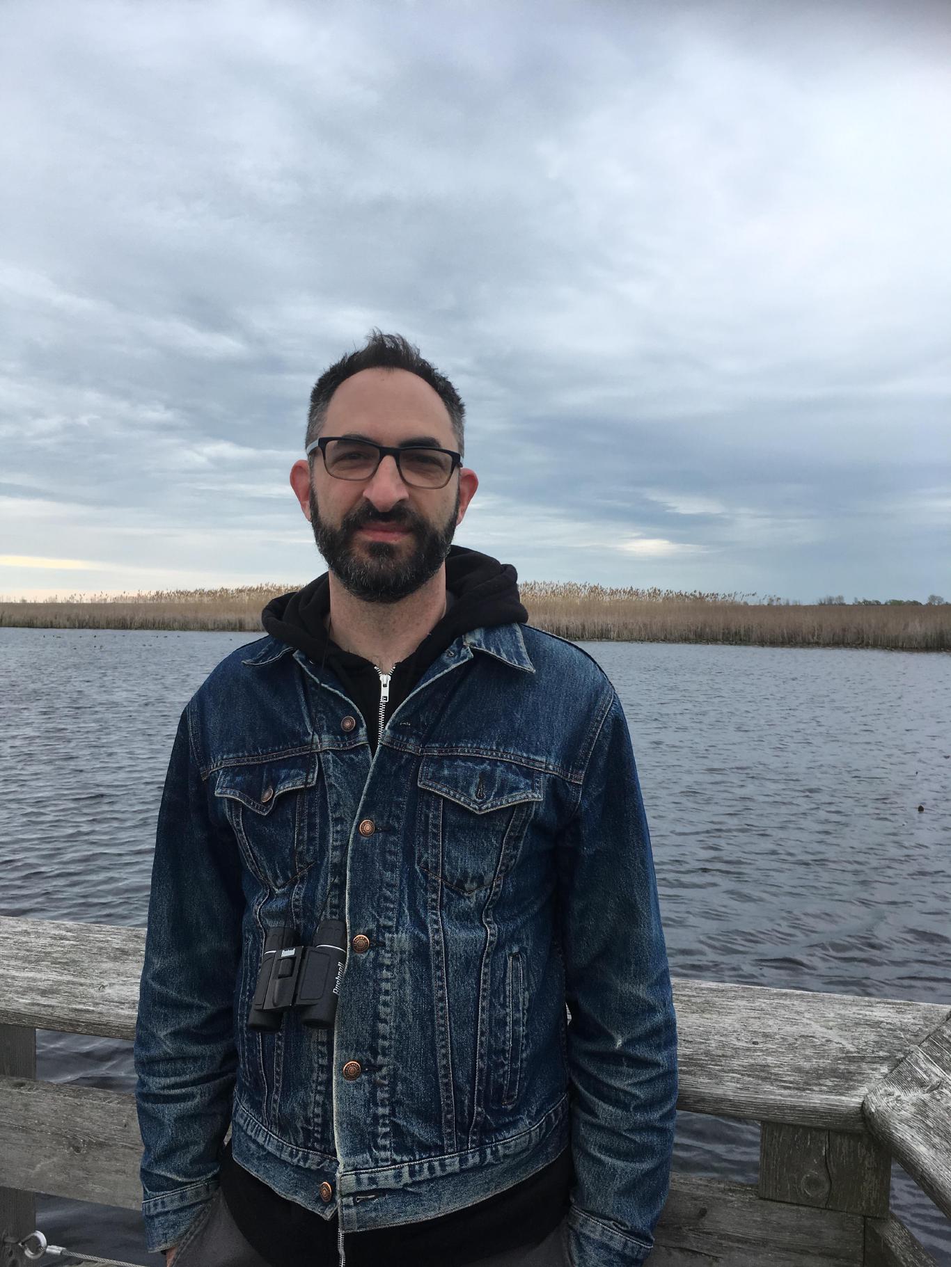 A photo of a bearded man with light skin tone in front of a choppy lake.